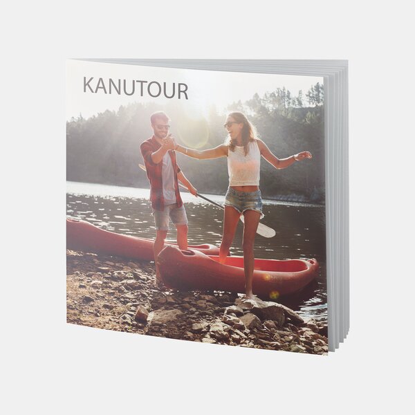 Ein quadratisches Fotobuch mit dem Titel „Kanutour“ zeigt ein junges Paar beim Einstieg ins Kanu am sonnigen Seeufer.